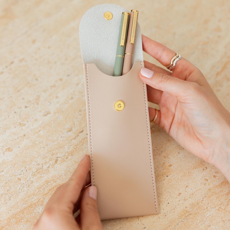 K&ouml;nnte beinhalten: Ein beiges Stifteetui mit goldenem Druckknopfverschluss enth&auml;lt zwei schlanke Stifte, einen in Salbeigr&uuml;n und einen in hellem Beige, beide mit goldenen Clips und Akzenten. Das Etui hat eine geschwungene Klappe und sichtbare Seitenn&auml;hte.