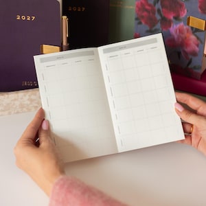 May include: An open meal planner notebook with a grid layout for breakfast, lunch, dinner, and snacks across seven days of the week. The pages are white with dark gray text. In the background, two dark purple 2027 planners and a floral planner.