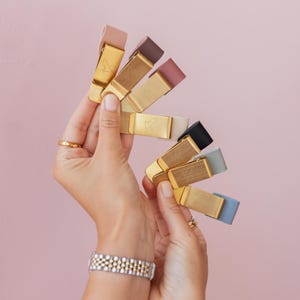 May include: A collection of money clips in various colors, including pink, brown, black, and blue. Each clip has a gold-colored metal base with a simple design. The clips are held in a person's hands against a pink background.