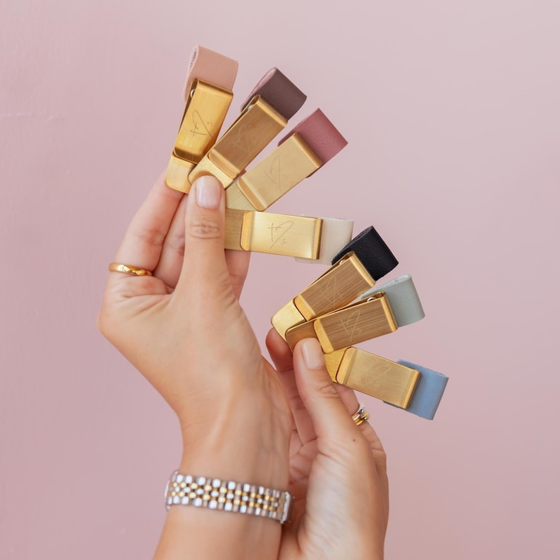 May include: A collection of money clips in various colors, including pink, brown, black, and blue. Each clip has a gold-colored metal base with a simple design. The clips are held in a person's hands against a pink background.