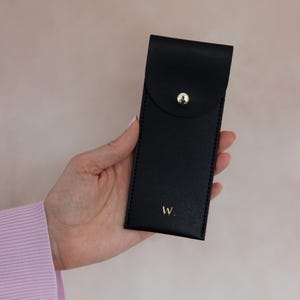 K&ouml;nnte beinhalten: Ein schwarzes, rechteckiges Etui mit abgerundeter Verschlussklappe, die durch einen goldfarbenen Metalldruckknopf gesichert ist. Auf der Vorderseite befindet sich der Buchstabe W mit einem Punkt in goldener Schrift f&uuml;r ein schlichtes Design.