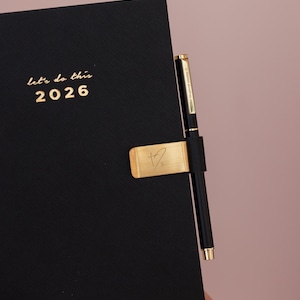 May include: Black 2026 planner with gold lettering that reads "let's do this". A gold pen holder with a signature and a black pen are attached to the planner. The background is a solid pink color.