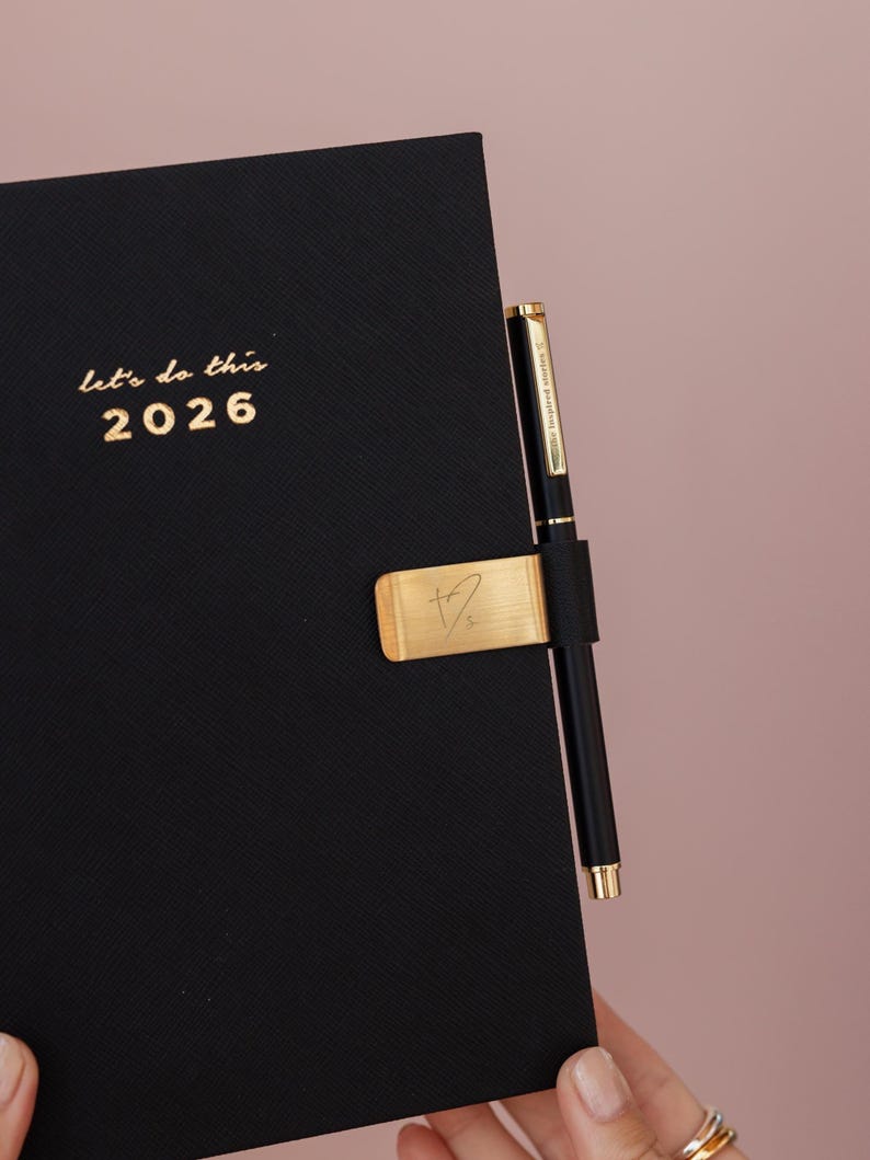 May include: Black 2026 planner with gold lettering that reads "let's do this". A gold pen holder with a signature and a black pen are attached to the planner. The background is a solid pink color.