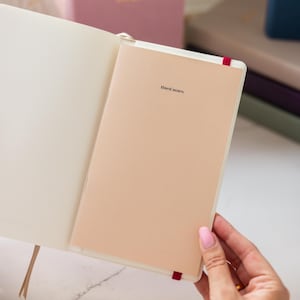 May include: An open notebook with a cream-colored cover featuring the text "lined notes" in black font. The notebook has a red elastic band closure and white interior pages. The design is simple, with a focus on the light tan paper cover and binding.