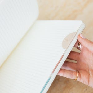 May include: Open notebook with lined pages. A beige, half-moon shaped bookmark is attached to the edge of the pages. The notebook is being held open by a person. The notebook's pages are white with thin gray lines.