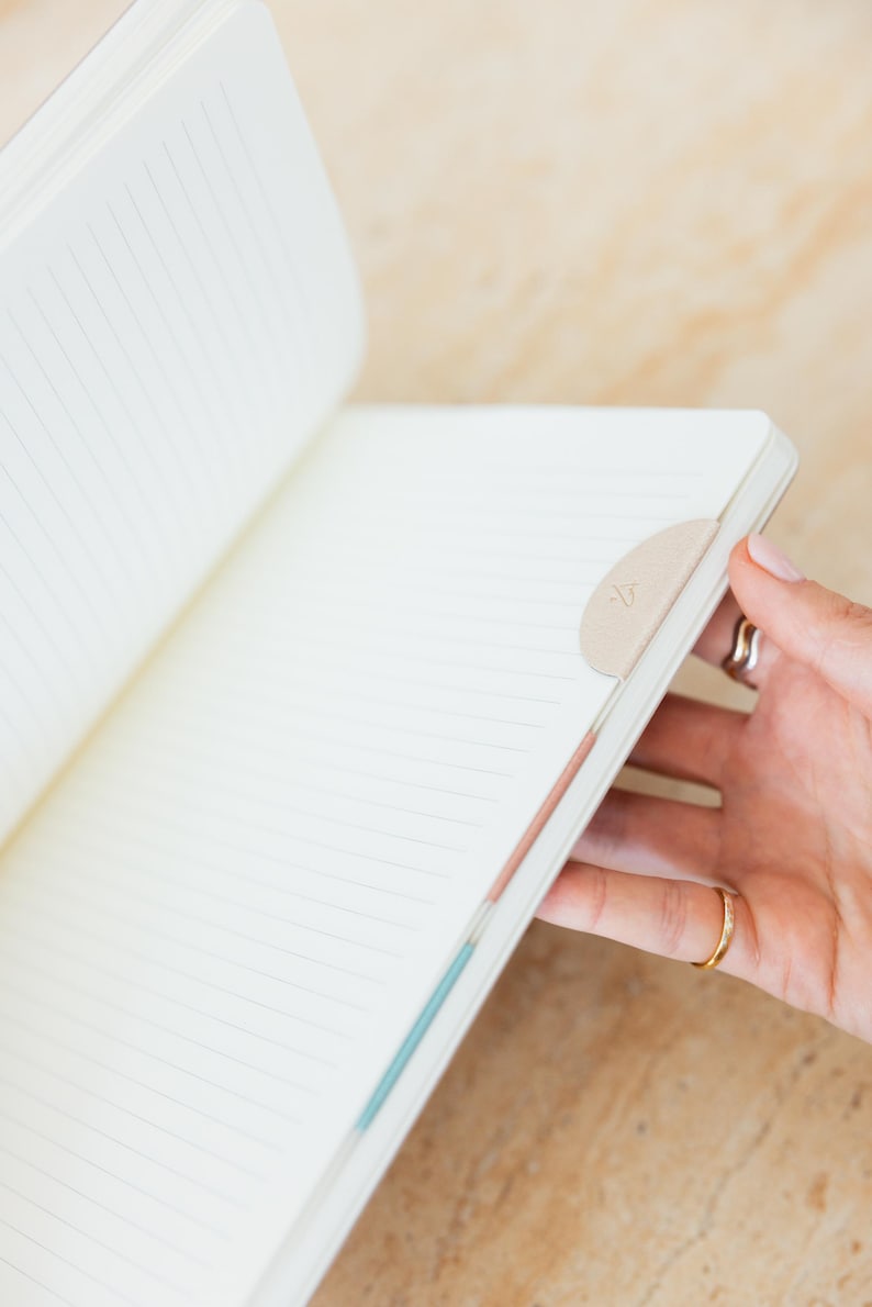 May include: Open notebook with lined pages. A beige, half-moon shaped bookmark is attached to the edge of the pages. The notebook is being held open by a person. The notebook's pages are white with thin gray lines.