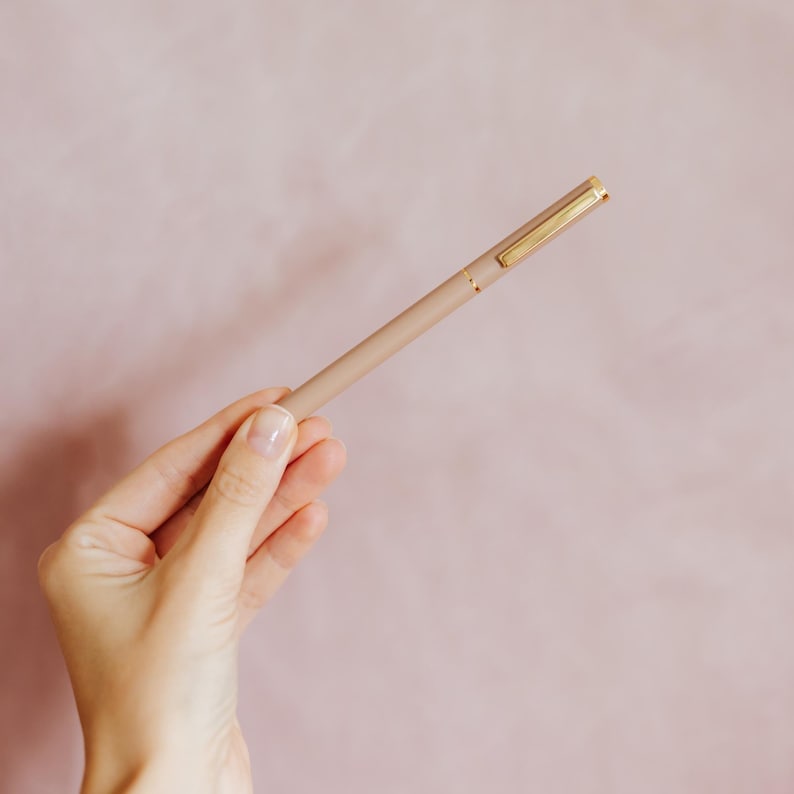 May include: A slim, matte beige ballpoint pen featuring a gold-toned metal clip and a thin gold ring accent near the cap. The pen has a smooth, cylindrical barrel design with a matching beige cap that fits securely onto the main body of the writing tool.
