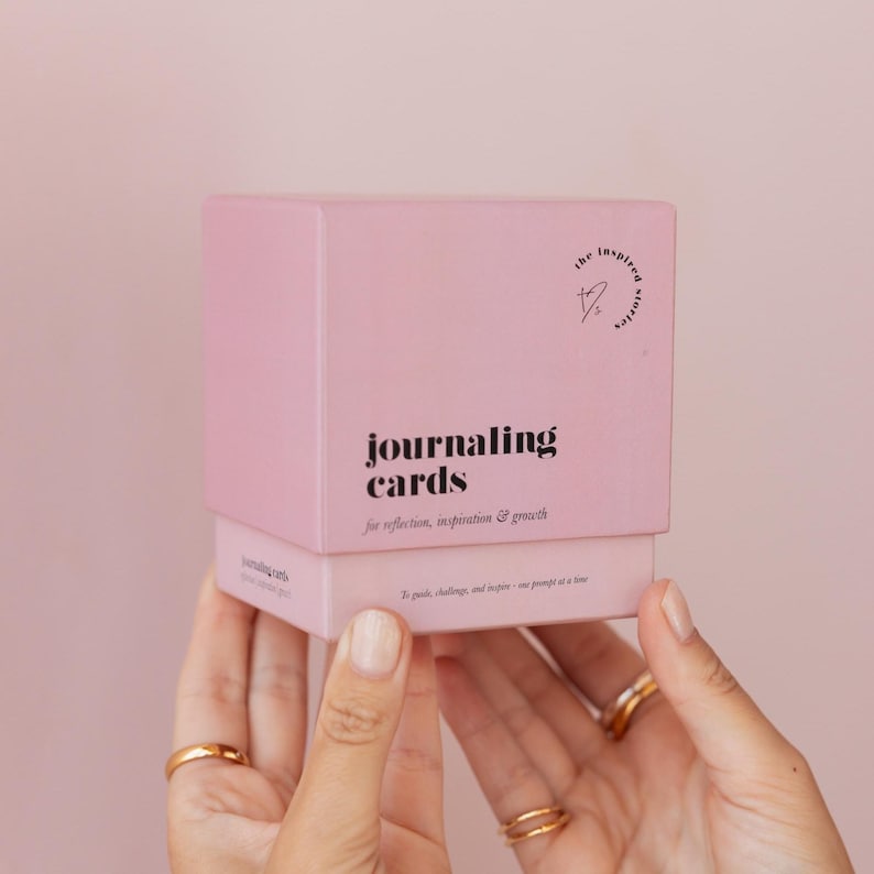 May include: A light pink box of journaling cards is held by two hands. The box has the words "journaling cards" in a bold, black font. The box also has the text "for reflection, inspiration & growth". The box has a logo that says "the inspired stories".