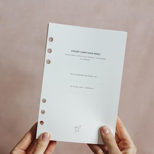 May include: A white undated weekly planner with the text "create your own story" and "UNDATED LIFESTYLE WEEKLY PLANNER 52 WEEKS". The planner has a section for "THIS PLANNER BELONGS TO" and "IN CASE LOST, CONTACT".