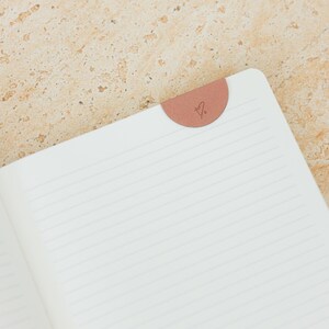 May include: Open notebook with lined pages and a salmon-colored bookmark. The bookmark is a half-circle shape with a small, delicate design. The notebook is open, revealing the white pages and the lines.