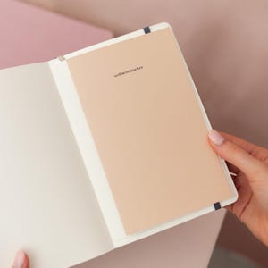 May include: An open notebook with a light beige page featuring the text wellness tracker printed in small black letters. The notebook has a white interior cover and a dark blue elastic band visible at the top and bottom edges of the page.