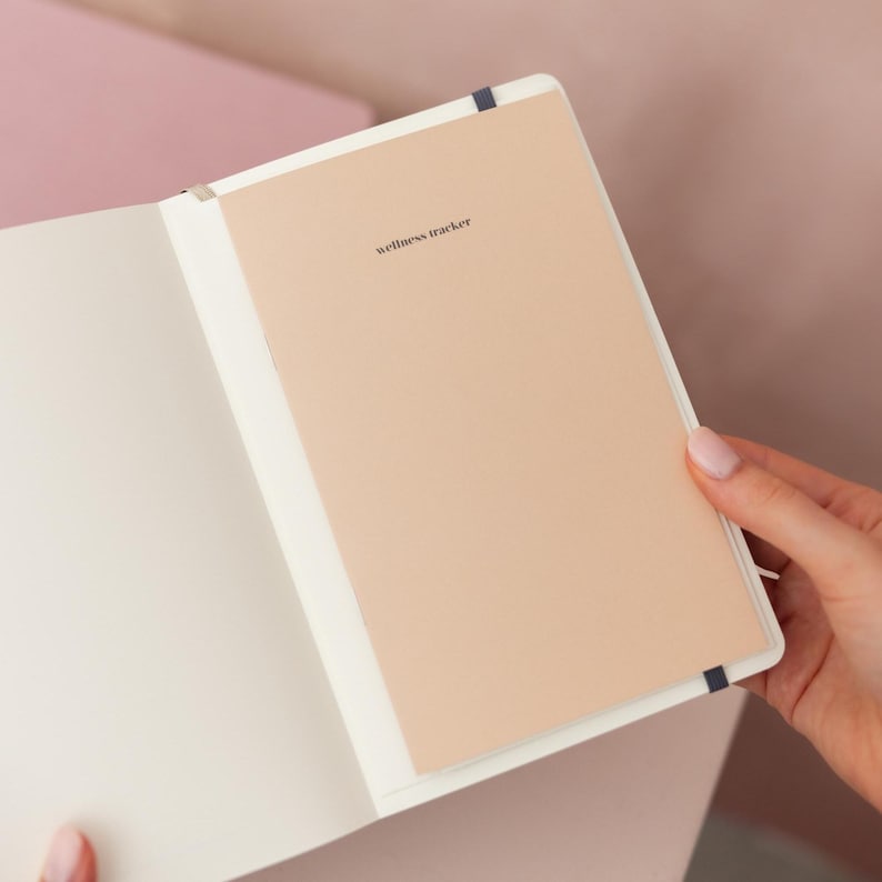 May include: An open notebook with a light beige page featuring the text wellness tracker printed in small black letters. The notebook has a white interior cover and a dark blue elastic band visible at the top and bottom edges of the page.