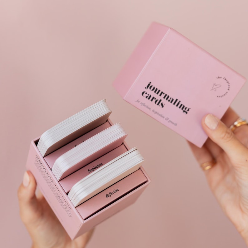 May include: A pink box set of journaling cards with the text "journaling cards" and "for reflection, inspiration & growth". The box is open, revealing several stacks of cards labeled "Inspiration" and "Reflection".