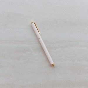 May include: A slim, cylindrical ballpoint pen featuring a matte off-white barrel and polished gold-tone metal accents. The design includes a gold pocket clip at the top and a matching gold tip at the base, providing a clean and functional writing tool.