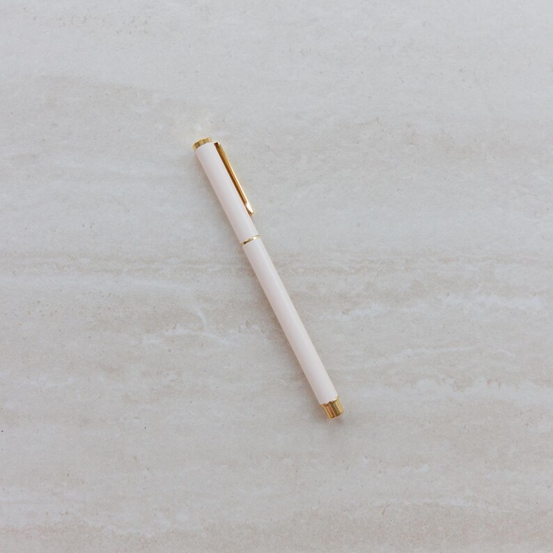 May include: A slim, cylindrical ballpoint pen featuring a matte off-white barrel and polished gold-tone metal accents. The design includes a gold pocket clip at the top and a matching gold tip at the base, providing a clean and functional writing tool.