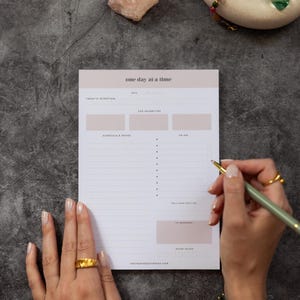 Può includere: Un blocco note bianco con la scritta "one day at a time" in alto. Il blocco note ha sezioni per le intenzioni quotidiane, le priorità principali, il programma, la lista delle cose da fare e la cura di sé. Una mano tiene una penna, pronta a scrivere.