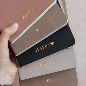 K&ouml;nnte beinhalten: Ein Stapel aus f&uuml;nf rechteckigen Notizb&uuml;chern mit matten Einb&auml;nden in den Farben Altrosa, Taupe, Schwarz, Hellgrau und Braun. Jedes Notizbuch ist mit goldener Folienpr&auml;gung versehen, darunter der Text KATO, ein Stern, HAPPY mit Herz, GEORGIE und 2026.