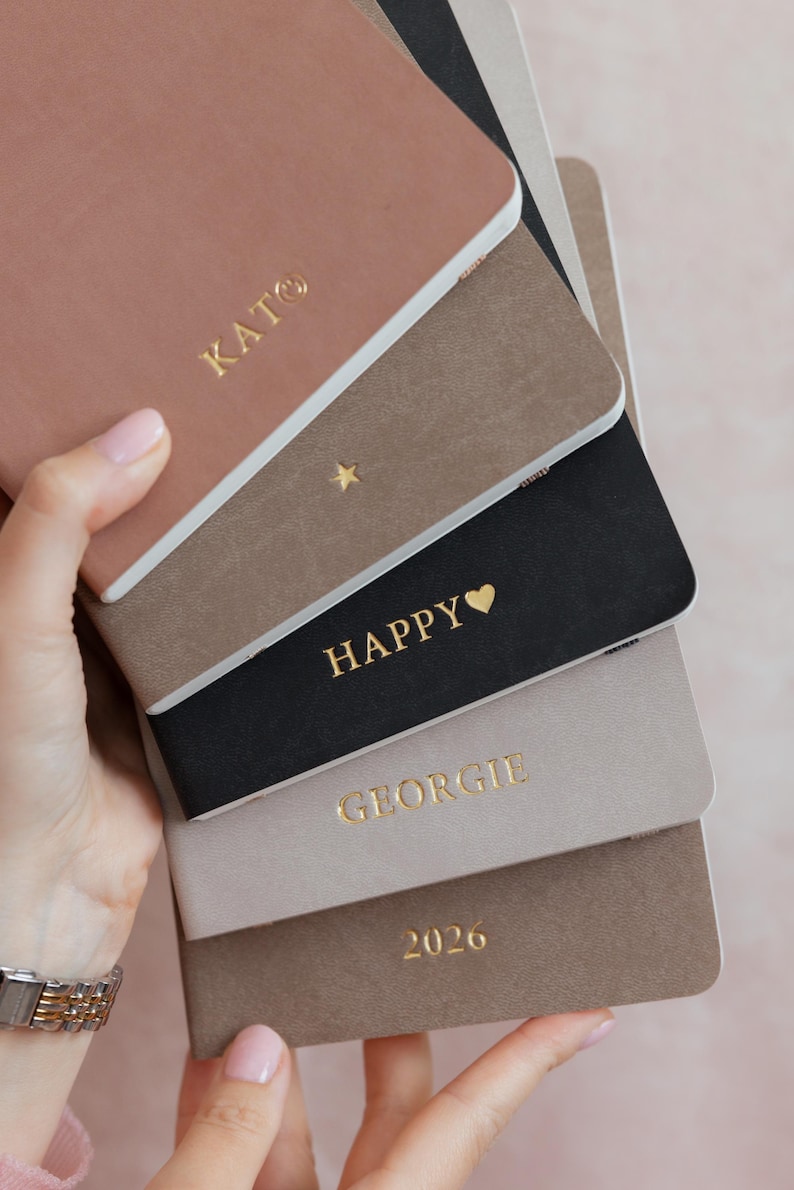 K&ouml;nnte beinhalten: Ein Stapel aus f&uuml;nf rechteckigen Notizb&uuml;chern mit matten Einb&auml;nden in den Farben Altrosa, Taupe, Schwarz, Hellgrau und Braun. Jedes Notizbuch ist mit goldener Folienpr&auml;gung versehen, darunter der Text KATO, ein Stern, HAPPY mit Herz, GEORGIE und 2026.