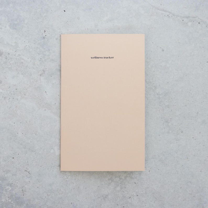 May include: A rectangular wellness tracker notebook with a solid light beige cover. The words wellness tracker are printed in small, black, lowercase sans-serif font centered near the top of the front cover. The notebook has a clean, flat design.