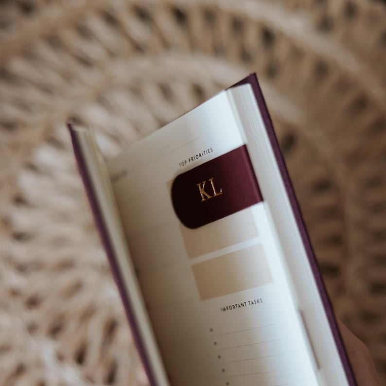 May include: A burgundy journal with a matching ribbon bookmark displaying the initials "KL" in gold. The open pages show sections labeled "Top Priorities" and "Important Tasks."