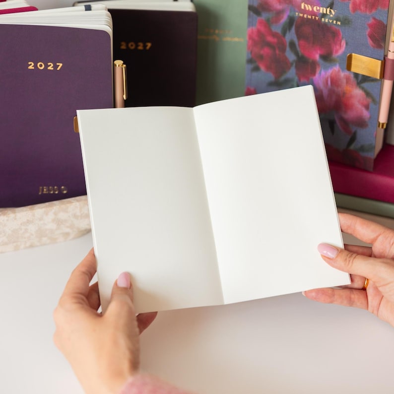 May include: An open notebook with blank white pages held in front of several closed planners. The planners include a dark purple one labeled 2027, a black one labeled 2027, and a floral patterned planner with the text twenty TWENTY SEVEN.