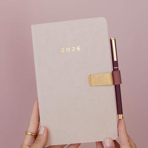 May include: A beige 2026 planner with a gold pen and pen holder. The pen is burgundy with gold accents. The planner has the year 2026 in gold lettering. The pen holder is gold with a heart-shaped design.
