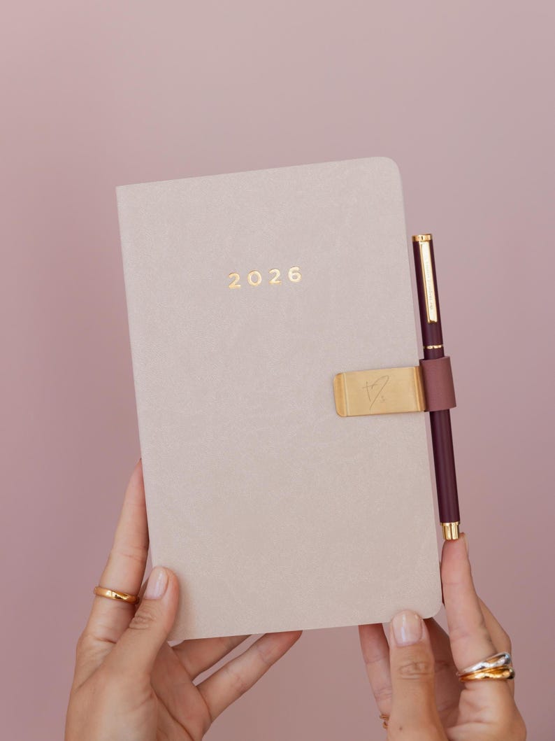 May include: A beige 2026 planner with a gold pen and pen holder. The pen is burgundy with gold accents. The planner has the year 2026 in gold lettering. The pen holder is gold with a heart-shaped design.