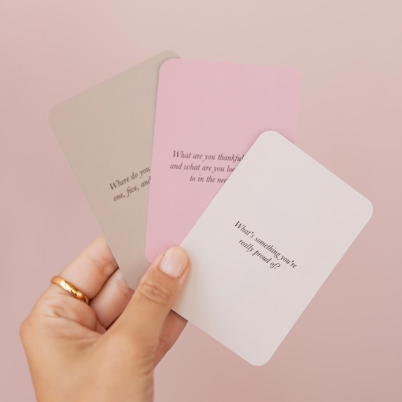 May include: Three pastel-colored cards with rounded corners are held in a hand. Each card has a different question printed on it in black. The cards are in shades of pink, beige, and white.