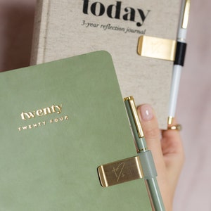 May include: A sage green journal with the gold text "twenty TWENTY FOUR" is held next to a journal with the text "today 3-year reflection journal". A gold pen clip holds a green pen. The journals are for reflection.