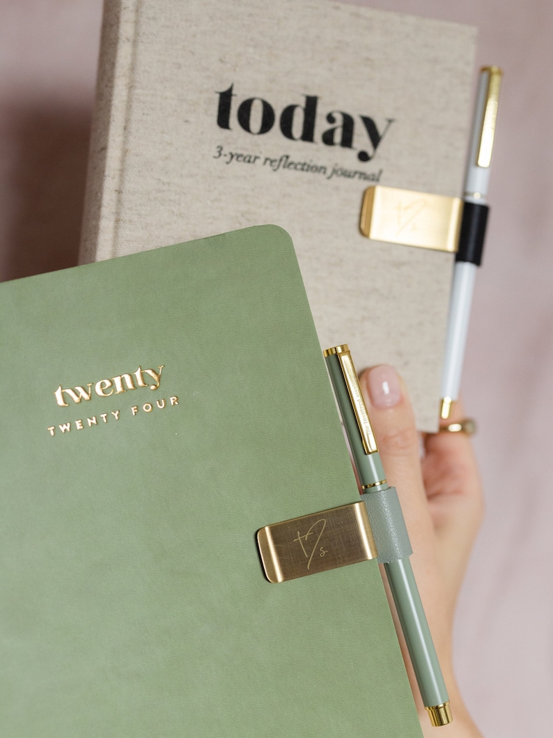 May include: A sage green journal with the gold text "twenty TWENTY FOUR" is held next to a journal with the text "today 3-year reflection journal". A gold pen clip holds a green pen. The journals are for reflection.