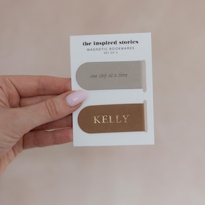 May include: A set of two magnetic bookmarks on a white card. One bookmark is tan with the phrase "one step at a time" printed on it. The other bookmark is brown with the name "KELLY" printed on it. The card reads "the inspired stories MAGNETIC BOOKMARKS SET OF 2".