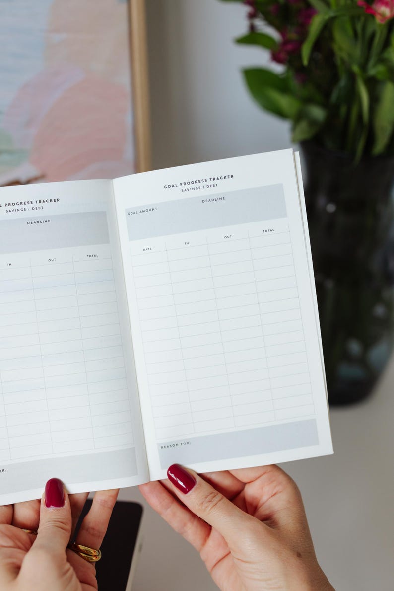 May include: An open planner with white pages featuring a Goal Progress Tracker for savings and debt. The layout includes columns for date, in, out, and total, with header sections for goal amount and deadline, and a bottom section labeled Reason For.