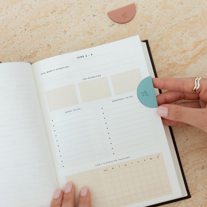 May include: Open planner with a teal bookmark and a pink bookmark on a light brown surface. The planner is open to a page with the text "JUNE 2-8" and sections for "TOP PRIORITIES" and "WORK TO-DO".