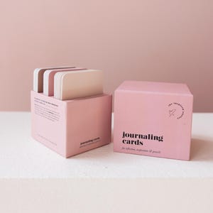 May include: Two pink boxes of journaling cards. One box is open, revealing a stack of cards with rounded corners. The other box is closed, with the words "journaling cards" printed on the front. The text "for reflection, inspiration & growth" is also printed on the box.
