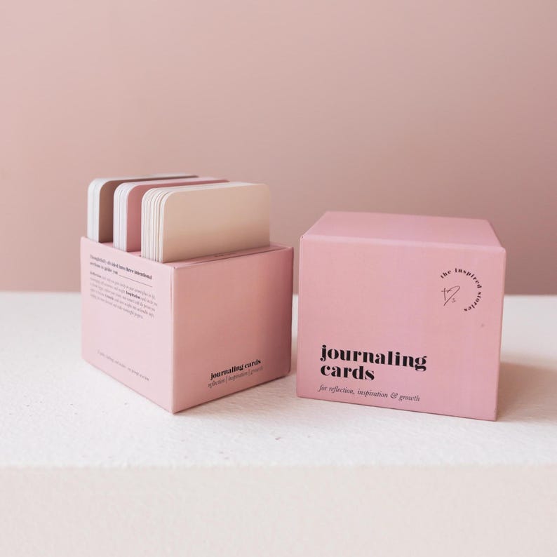 May include: Two pink boxes of journaling cards. One box is open, revealing a stack of cards with rounded corners. The other box is closed, with the words "journaling cards" printed on the front. The text "for reflection, inspiration & growth" is also printed on the box.