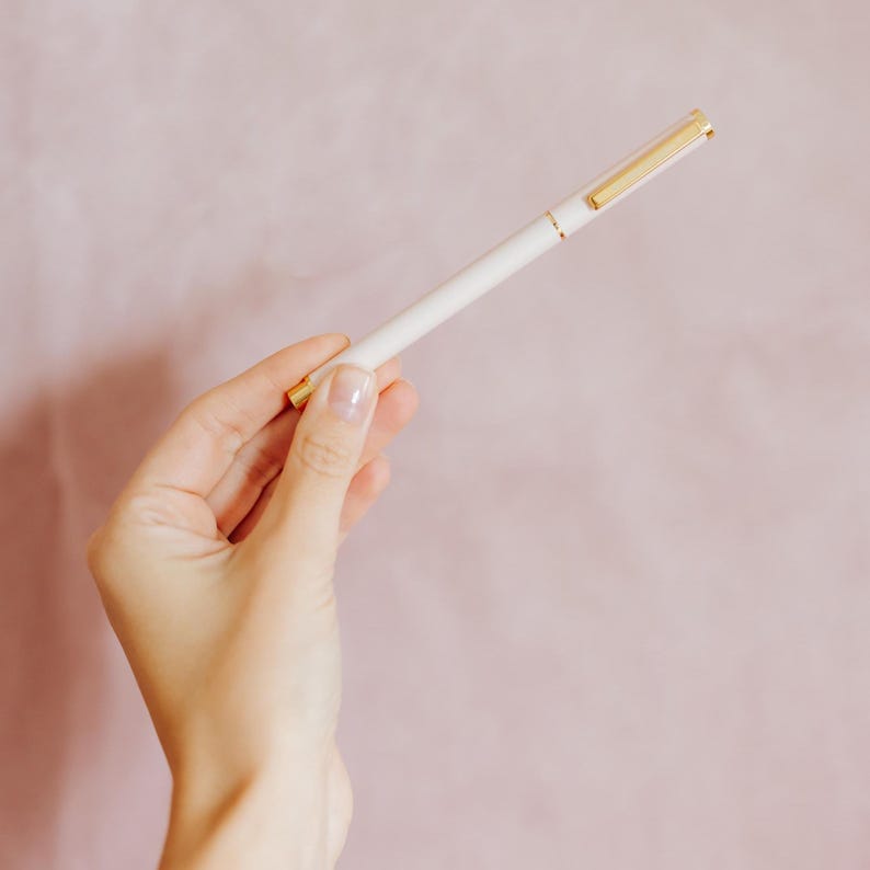 May include: A slim white ballpoint pen featuring a polished gold-tone clip and matching gold-tone accents at the tip and cap base. The pen has a smooth, cylindrical barrel design with a clean finish, providing a simple and functional writing instrument.