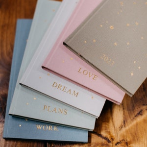Puede incluir: Cinco cuadernos de tapa blanda apilados con acentos de estrellas doradas. El cuaderno superior es gris y dice "2023". El siguiente cuaderno es rosa y dice "LOVE". El siguiente cuaderno es blanco y dice "DREAM". El siguiente cuaderno es azul claro y dice "PLANS". El cuaderno inferior es azul y dice "WORK".