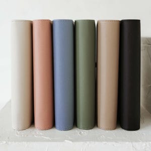 Peut inclure: Sept carnets reliés en cuir de différentes couleurs alignés en rangée. Les couleurs sont beige, rose clair, bleu clair, vert olive, beige et noir.