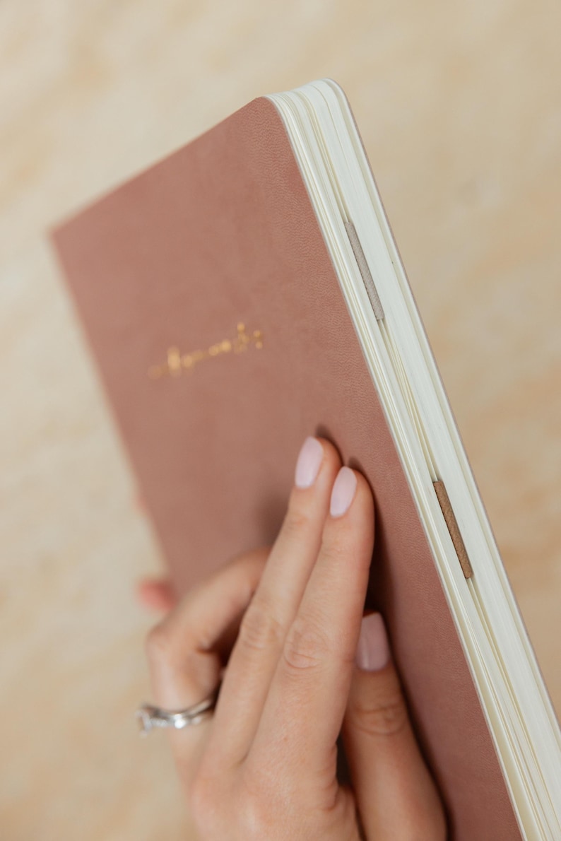 May include: A dusty rose-colored notebook with gold foil lettering. The notebook has a soft cover and cream-colored pages. The binding is visible, with a few small, tan-colored strips holding the pages together.