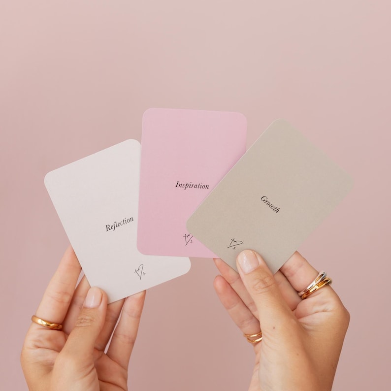 May include: Three pastel-colored cards with the words "Reflection", "Inspiration", and "Growth" printed on them. The cards are held in front of a pink background. Each card has a small symbol at the bottom.