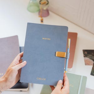 Op de afbeelding: Een leiblauw notitieboek met een gouden metalen penhouder en een bijpassende pen. De kaft is voorzien van het woord NOTES in gouden letters aan de bovenkant en de naam CHLOE in gouden letters aan de onderkant. Het notitieboek heeft een matte afwerking.
