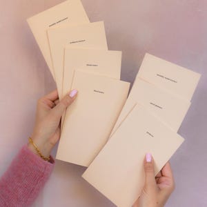 May include: A collection of light beige paper booklets with black text. The booklets are labelled monthly habit tracker, wellness tracker, blank notes, lined notes, meal planner, and projects. Each booklet has a simple, clean design with text.