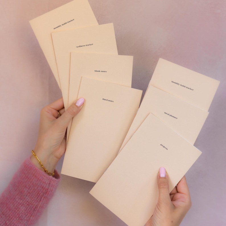 May include: A collection of light beige paper booklets with black text. The booklets are labeled monthly habit tracker, wellness tracker, blank notes, lined notes, meal planner, and projects. Each booklet has a simple, clean design with text.