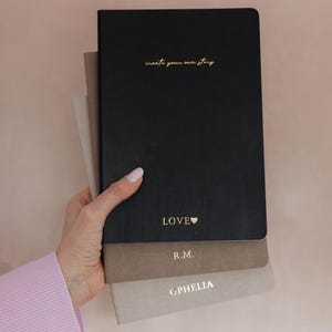 Peut inclure: Une pile de trois carnets rectangulaires aux couvertures mates en noir, taupe et beige clair. Le carnet noir porte le texte doré create your own story et LOVE avec un cœur. Le carnet taupe affiche R.M. et le carnet beige porte le nom OPHELIA.