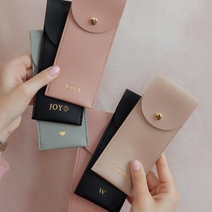 K&ouml;nnte beinhalten: Eine Sammlung rechteckiger Etuis in Schwarz, Altrosa und Salbeigr&uuml;n, jeweils mit einem goldenen Druckknopfverschluss. Die Etuis sind mit goldenem Text personalisiert, darunter LISA, JOY, MUM, W und K&A, sowie einem kleinen Herz- und Smiley-Symbol.