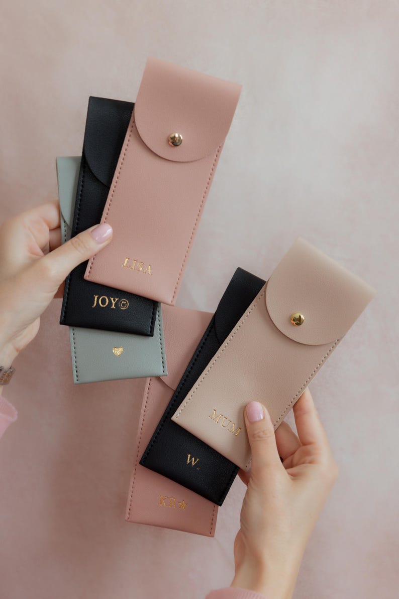 K&ouml;nnte beinhalten: Eine Sammlung rechteckiger Etuis in Schwarz, Altrosa und Salbeigr&uuml;n, jeweils mit einem goldenen Druckknopfverschluss. Die Etuis sind mit goldenem Text personalisiert, darunter LISA, JOY, MUM, W und K&A, sowie einem kleinen Herz- und Smiley-Symbol.