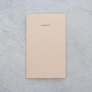 May include: A rectangular meal planner notebook with a solid light beige cover. The words meal planner are printed in a small, black, sans-serif font centered near the top of the front cover. The notebook has a clean, flat, and smooth surface.