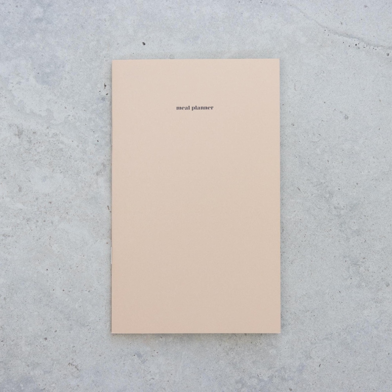 May include: A rectangular meal planner notebook with a solid light beige cover. The words meal planner are printed in a small, black, sans-serif font centered near the top of the front cover. The notebook has a clean, flat, and smooth surface.