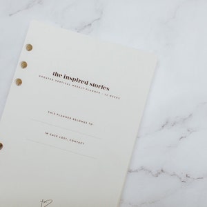 May include: A white planner with the text "the inspired stories" in brown lettering. The planner is a vertical weekly planner with 52 weeks. The text "This planner belongs to" and "In case lost, contact" are printed on the planner.