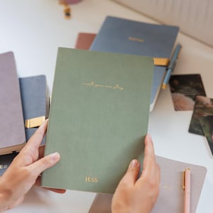 Könnte beinhalten: Ein salbeigrünes Notizbuch mit goldfarbenem Text, der create your own story und JESS lautet. Das Notizbuch wird vor anderen Notizbüchern in Blau-, Lila- und Rosatönen gehalten, zusammen mit einem rosa Stift und kleinen Karten auf einer weißen Fläche.